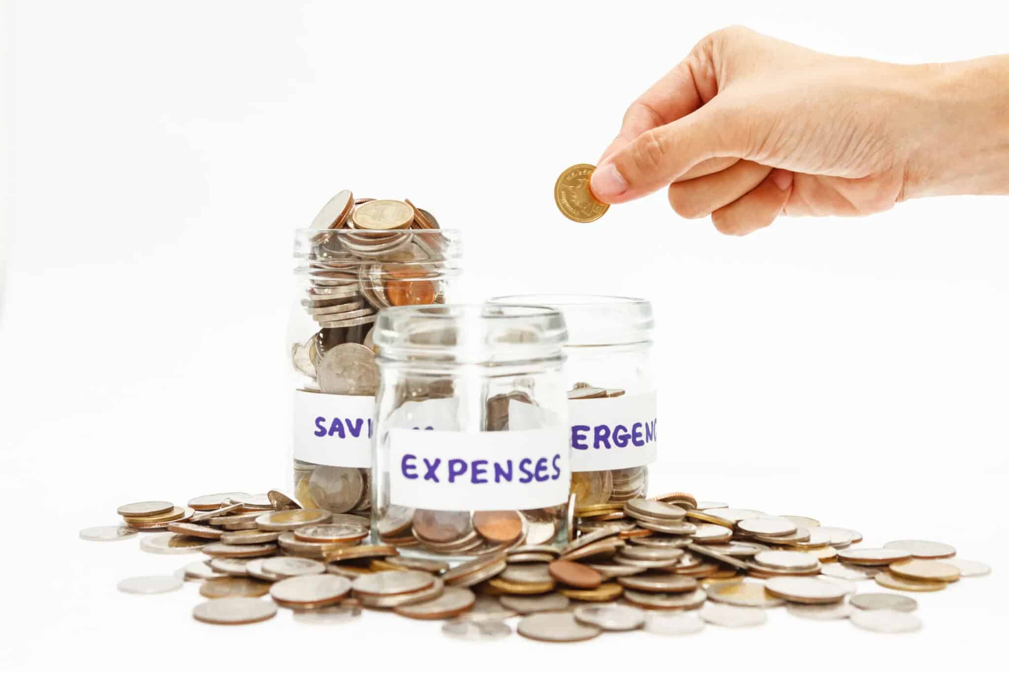 Hand placing a coin into a labeled jar for emergency savings, surrounded by additional jars for expenses and savings, illustrating financial management and budgeting strategies.